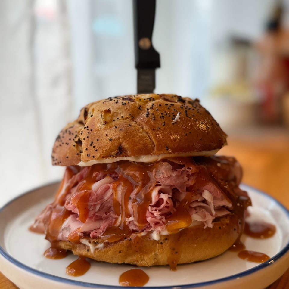 Overstuffed roast beef sandwich on a seeded bun, layered with melted cheese and brown sauce on a plate.