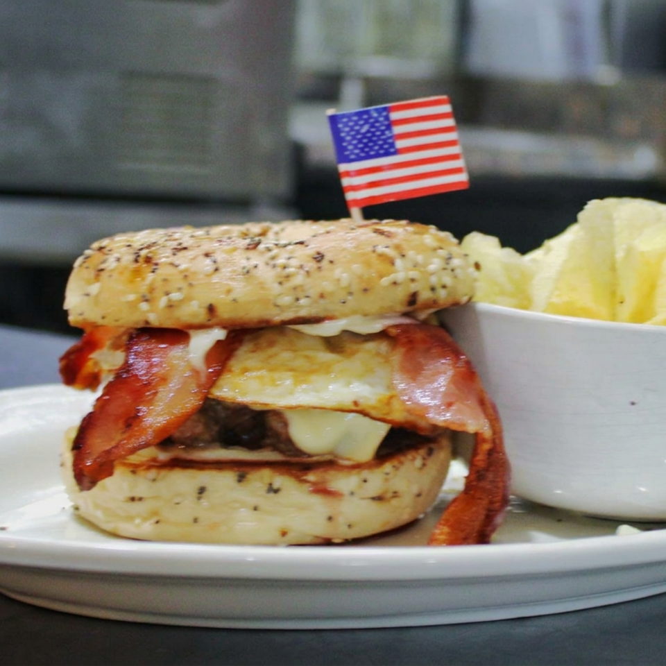 Bacon cheeseburger on a seasoned bun with potato chips, served on a white plate and topped with an American flag pick.